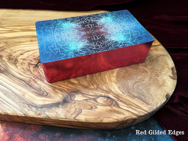 May include: A rectangular deck of cards featuring a blue and purple cosmic-themed back design with intricate white geometric patterns. The sides of the card deck are finished with a metallic red foil, creating distinct red gilded edges.