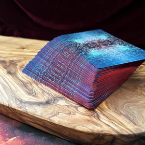 May include: A deck of rectangular playing cards with a blue and teal cosmic design on the back. The edges of the cards are colored in a vibrant red, creating a distinct contrast against the dark blue patterned surface of the top card.
