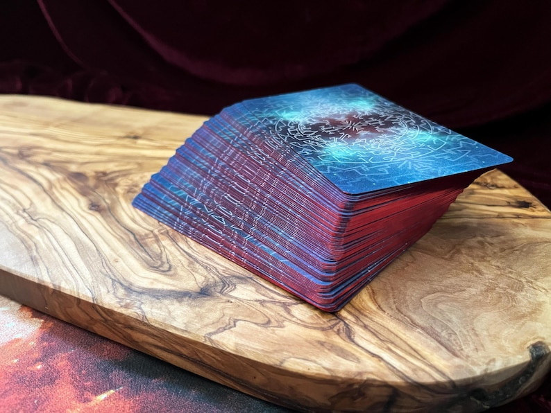 May include: A deck of rectangular playing cards with a blue and teal cosmic design on the back. The edges of the cards are colored in a vibrant red, creating a distinct contrast against the dark blue patterned surface of the top card.