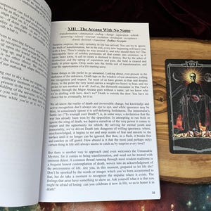 May include: An open book with text titled XIII - The Arcana With No Name and a tarot card featuring a skeletal figure in a black robe, a glowing sun, a crescent moon, and a pile of skulls at the base, set against a dark, starry background.