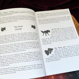 May include: An open book with white pages and black text titled The Fool's Journey. The left page discusses the Major Arcana, while the right page details the Minor Arcana, specifically the Fire suit with a wolf icon and the Water suit with a fish icon.