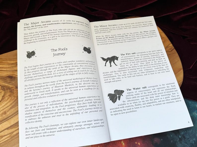 May include: An open book with white pages and black text titled The Fool's Journey. The left page discusses the Major Arcana, while the right page details the Minor Arcana, specifically the Fire suit with a wolf icon and the Water suit with a fish icon.