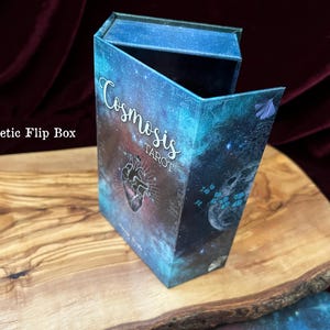 May include: A rectangular magnetic flip box featuring a deep blue and purple galaxy print. The front displays the text "Cosmosis TAROT" in white script and block lettering, accompanied by a central anatomical heart illustration and small blue butterflies.
