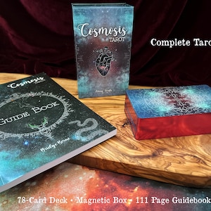 May include: A Cosmosis Tarot set featuring a 78-card deck with red metallic edges, a magnetic storage box, and a 111-page guidebook. The box and book covers display a dark blue and teal nebula design with a central anatomical heart illustration.