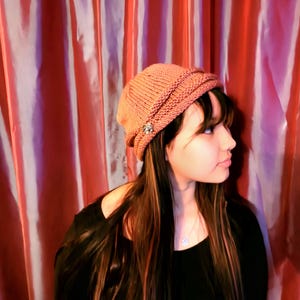 May include: A close-up of a person wearing a pink knitted beanie with a decorative button. The beanie is folded down at the brim and the person is looking to the side. The background is a red and white striped curtain.