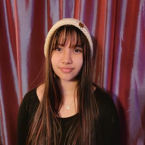 May include: A young person with long brown hair wearing a white knit beanie with a brown button on top. They are wearing a black shirt and a silver necklace with a small pendant.