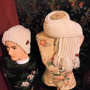 May include: Cream-colored knitted beanie hat and scarf set. The hat is on a mannequin head, and the scarf is draped over a floral dress form. Both items have a small brown leather tag.