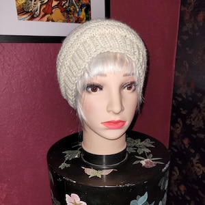 May include: A cream-colored, knitted beanie hat. The hat is displayed on a mannequin head with white hair, red lips, and dark eyelashes. The mannequin is placed on a black decorative stand with floral designs.