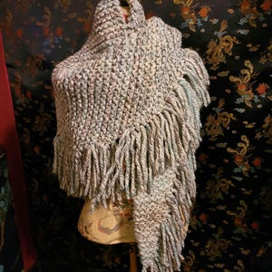 May include: A hand-knitted triangular shawl featuring a variegated yarn blend of light blue, cream, and grey tones. The garment has a textured garter stitch pattern and is finished with long, thick fringe tassels along the bottom edges.