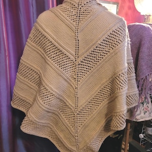 May include: A beige knitted shawl featuring a chevron pattern with alternating sections of stockinette stitch and openwork eyelet rows. The garment has a central vertical seam and a pointed hemline, showcasing a textured design throughout the piece.