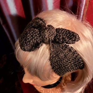 May include: A dark gray knitted headband with a large bow. The headband is worn on a mannequin with blonde hair.