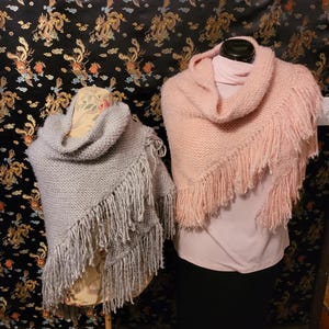 May include: Two triangular knit shawls, one in light gray and one in soft pink, each featuring a textured garter stitch pattern and long, dangling fringe along the diagonal edge. Both shawls are draped over display forms.
