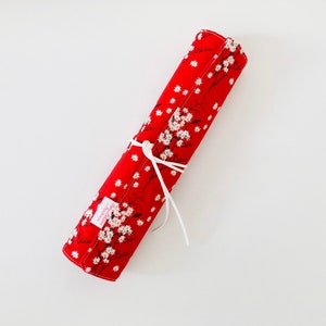 Peut inclure: Une trousse à crayons en tissu rouge avec un motif floral blanc et un ruban blanc noué autour.