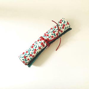 Peut inclure: Un étui à crayons en tissu roulé avec un fond blanc et un motif floral rouge et vert. L'étui est attaché avec une ficelle rouge.