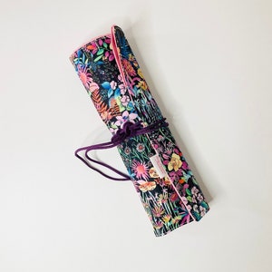 Peut inclure: Un étui à crayons en tissu noir roulé avec un imprimé floral rose, bleu, jaune et vert. L'étui est attaché avec un ruban violet et a une couture rose.
