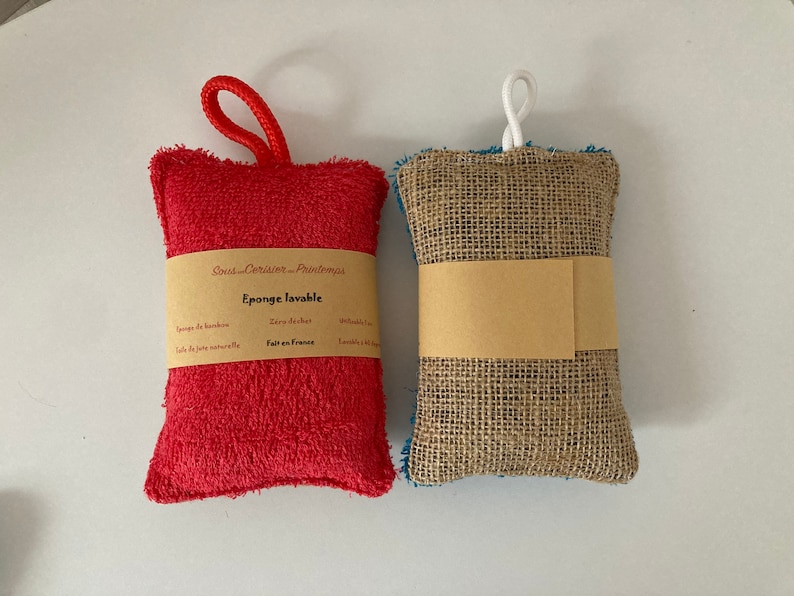 May include: Two reusable cleaning sponges. One is red and made of terry cloth with a loop handle. The other is brown and made of burlap with a white string handle. Both have labels that say "Sous-Cuisson - Printemps" and "Eponge lavable".