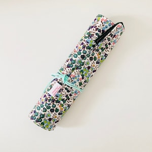 Puede incluir: Un estuche de lápices de tela enrollado con un patrón floral en tonos verdes, azules y rosas. El estuche está atado con una cinta azul claro y tiene un ribete verde oscuro.