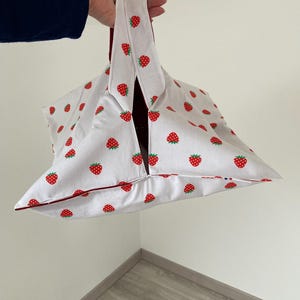 Può includere: Borsa tote bianca con un motivo ripetuto di fragole rosse. La borsa ha finiture e manici rosso scuro. La borsa è tenuta da una mano.