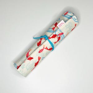 Peut inclure: Un rouleau de crayons en tissu blanc avec un motif de vagues bleu et blanc et un imprimé de poissons rouges. Le rouleau est attaché avec un ruban bleu et porte une étiquette rose qui dit "Sew Crafty Mama".