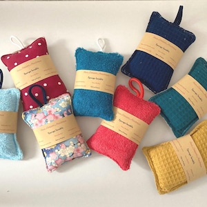May include: Eight colourful, rectangular, handmade bath sponges with a loop for hanging. Each sponge has a label that reads "Sponge scrubbie" and is made of different materials, including terry cloth, waffle weave, and patterned fabric.