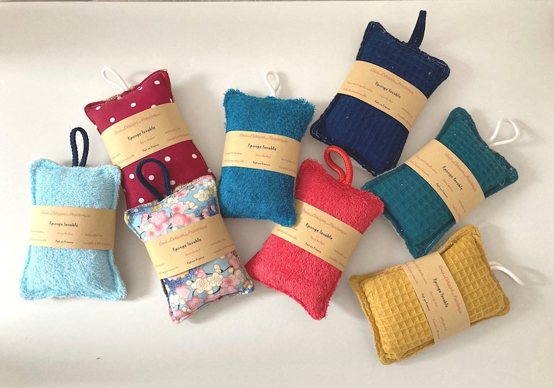 May include: Eight colourful, rectangular, handmade bath sponges with a loop for hanging. Each sponge has a label that reads "Sponge scrubbie" and is made of different materials, including terry cloth, waffle weave, and patterned fabric.