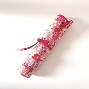 Peut inclure: Étui en tissu floral enroulé, avec un motif floral rose et une attache rose assortie. L'étui est probablement destiné à ranger des fournitures artistiques ou d'autres petits objets. Le tissu a un fond blanc avec des fleurs roses et rouges.