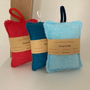 May include: Three colourful bath sponges with a brown label that reads "Sous-Coraline - Paris" and "Eponge lavable" in French. The sponges are made of a soft, textured material and are designed for washing dishes or cleaning.