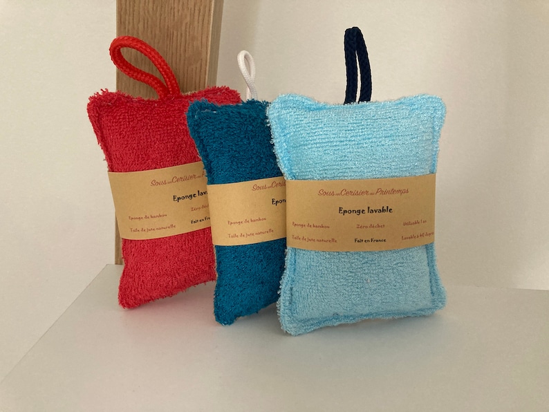 May include: Three colourful bath sponges with a brown label that reads "Sous-Coraline - Paris" and "Eponge lavable" in French. The sponges are made of a soft, textured material and are designed for washing dishes or cleaning.