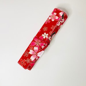 Può includere: Un rotolo di tessuto rosso con un motivo floreale bianco e rosa. Il rotolo è legato con uno spago rosso e ha un piccolo cartellino bianco con del testo.