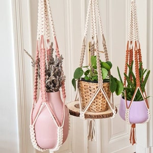 Puede incluir: Tres colgadores de plantas de macramé con aros de madera y borlas decorativas. Uno sostiene un jarrón rosa con flores secas, otro una cesta tejida sobre una repisa de madera y el tercero una maceta morada con una planta. Los colgadores cuelgan de una barra blanca.