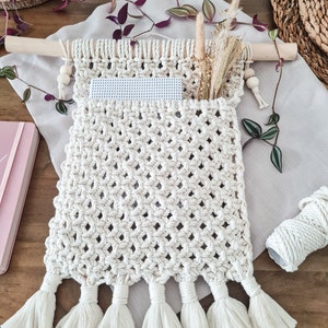 May include: A white macrame wall hanging with a pocket and tassels. The hanging is made with natural cotton rope and has a wooden dowel at the top.