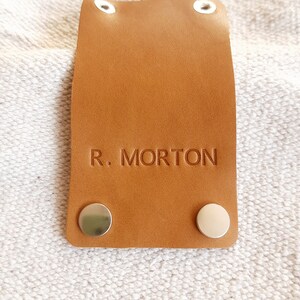 May include: A rectangular, tan leather tag with the embossed text "R. MORTON". The tag has two silver rivets at the bottom and two at the top. The tag is on a textured, off-white surface.