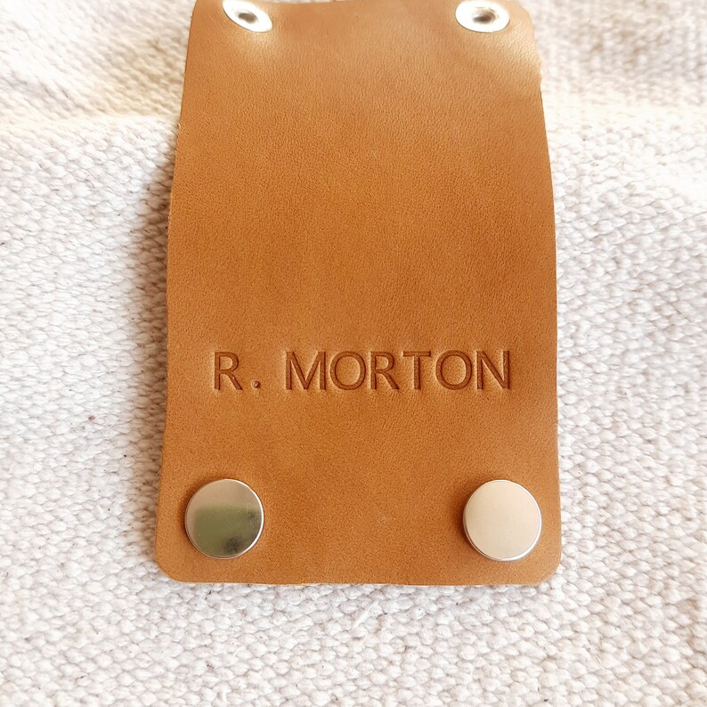 May include: A rectangular, tan leather tag with the embossed text "R. MORTON". The tag has two silver rivets at the bottom and two at the top. The tag is on a textured, off-white surface.