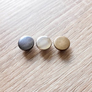 May include: Three round metal buttons in different finishes: dark gray, silver, and antique brass. The buttons are arranged in a row on a light wood-grain surface. These buttons could be used for clothing or craft projects.