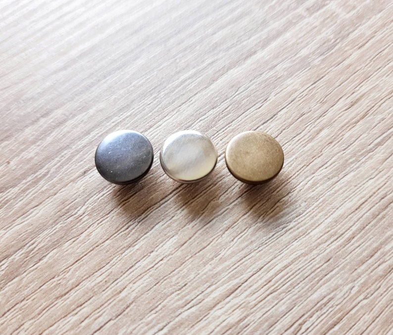 May include: Three round metal buttons in different finishes: dark gray, silver, and antique brass. The buttons are arranged in a row on a light wood-grain surface. These buttons could be used for clothing or craft projects.