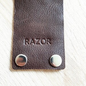 May include: Close-up of a dark brown leather strap with the word "RAZOR" embossed on it. Two silver metal snaps are visible at the bottom of the strap. The leather has a textured surface.