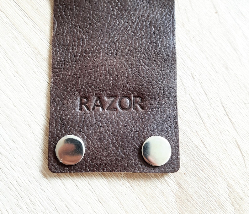 May include: Close-up of a dark brown leather strap with the word "RAZOR" embossed on it. Two silver metal snaps are visible at the bottom of the strap. The leather has a textured surface.