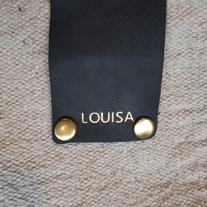May include: Close-up of a black leather strap with the gold-colored word "LOUISA" printed on it. Two gold-colored rivets are on the strap. The background is a textured, off-white fabric.