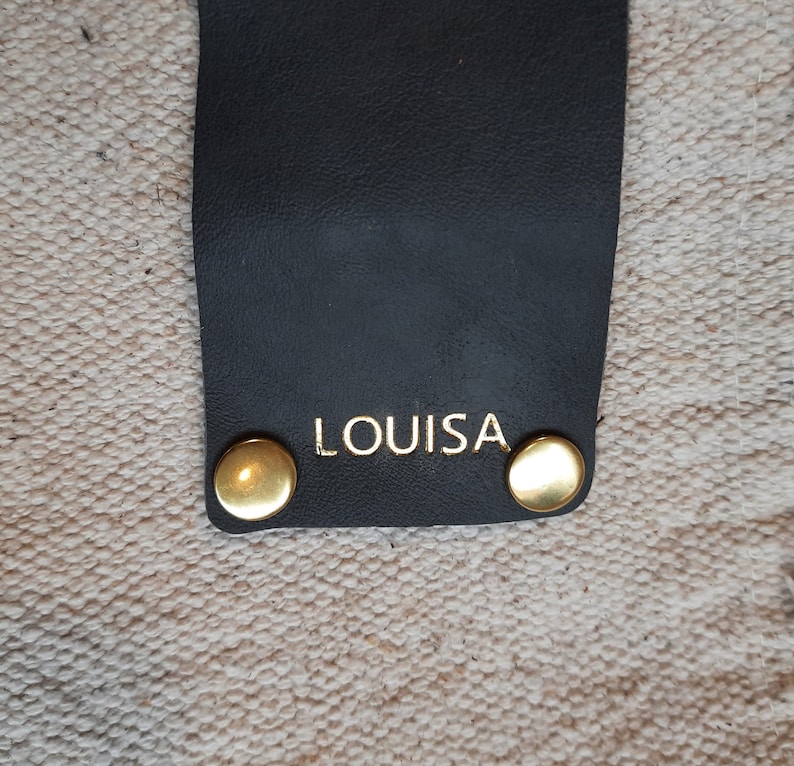 May include: Close-up of a black leather strap with the gold-colored word "LOUISA" printed on it. Two gold-colored rivets are on the strap. The background is a textured, off-white fabric.