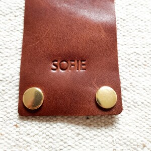 May include: Close-up of a brown leather tag with the name "SOFIE" embossed on it. Two gold-colored metal rivets are visible at the bottom. The tag is rectangular and appears to be made of high-quality leather.