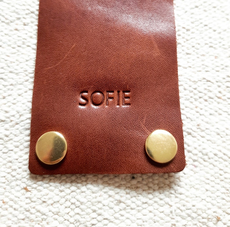 May include: Close-up of a brown leather tag with the name "SOFIE" embossed on it. Two gold-colored metal rivets are visible at the bottom. The tag is rectangular and appears to be made of high-quality leather.