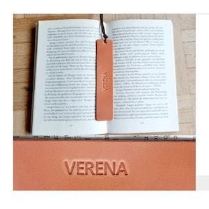 May include: A close-up of a brown leather bookmark with the name "VERENA" embossed in gold lettering. The bookmark is lying on top of an open book.