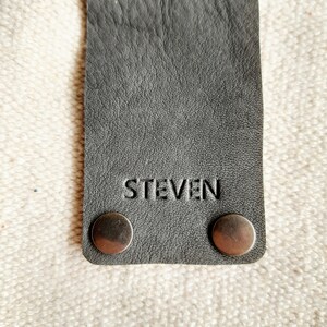 May include: A gray leather strap with the name "STEVEN" embossed on it. The strap has two silver rivets at the top and two at the bottom. The background is a textured, off-white fabric.