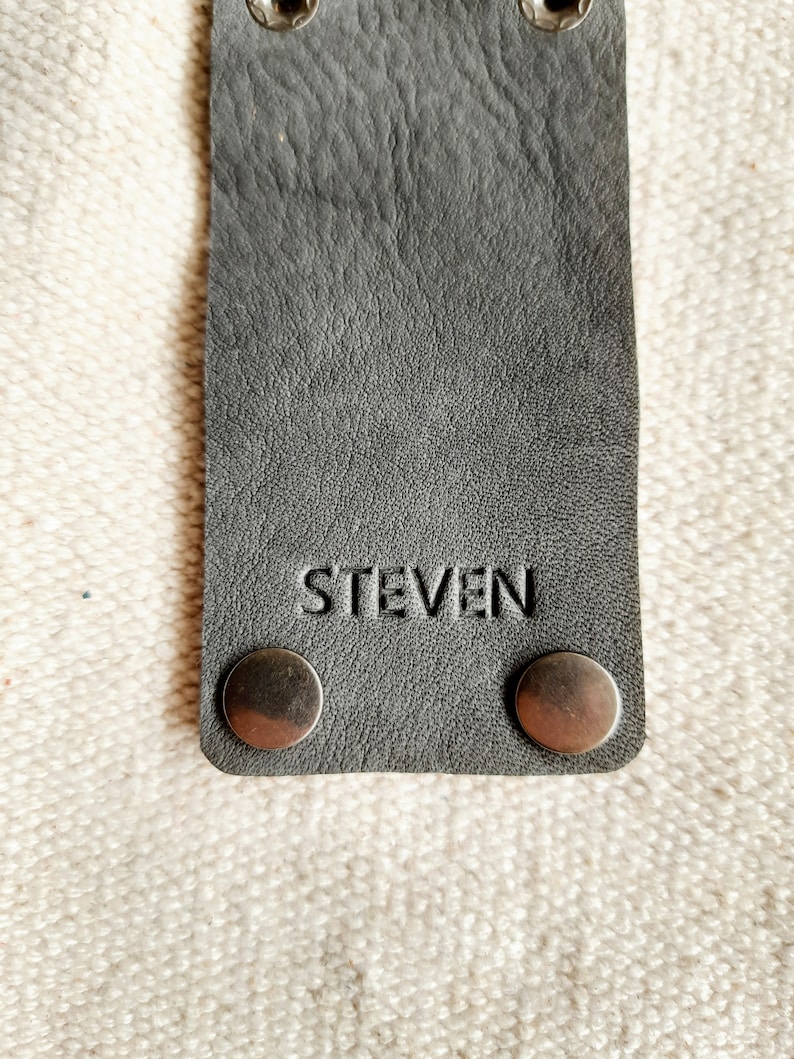May include: A gray leather strap with the name "STEVEN" embossed on it. The strap has two silver rivets at the top and two at the bottom. The background is a textured, off-white fabric.