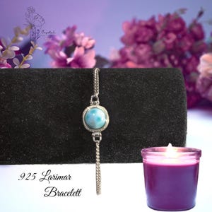 May include: A silver bracelet featuring a round, light blue, marbled Larimar gemstone set in a circular frame. The bracelet has a delicate chain and is displayed on a black velvet stand. The text .925 Larimar Bracelet is visible.