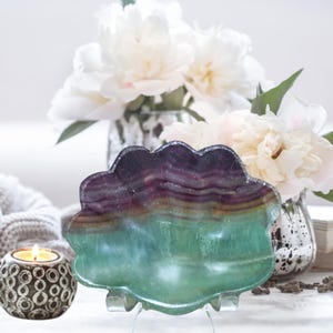 May include: A decorative, scalloped-edge fluorite dish in shades of purple, green, and yellow. The dish is displayed on a clear stand, with a lit candle and white flowers in the background. The fluorite dish is a unique home decor item.