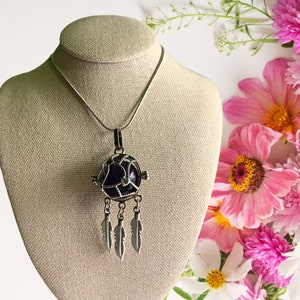 May include: A silver necklace with a purple and silver dreamcatcher pendant with feathers. The necklace is displayed on a beige mannequin.