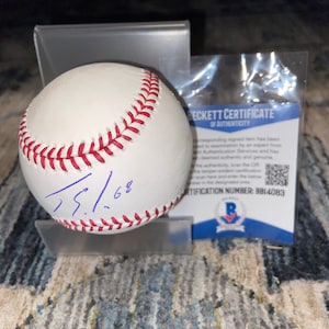 Puede incluir: Una pelota de béisbol blanca con costuras rojas y una firma azul. La pelota se exhibe en un soporte transparente, acompañada de un Certificado de Autenticidad Beckett. El certificado incluye un código QR y el número de certificación 8814083.