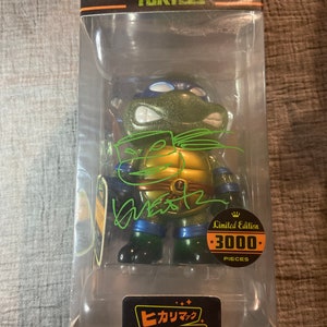May include: A Teenage Mutant Ninja Turtle figure in a clear plastic display case. The figure is green, blue, and gold, with a signature in green ink. The packaging includes the Nickelodeon and TMNT logos, and a sticker that says "Limited Edition 3000 Pieces."