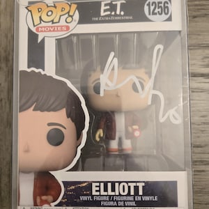 May include: A Funko Pop! Movies vinyl figure of Elliott from E.T. The Extra-Terrestrial, number 1256, in a clear plastic display case. The figure is wearing a red jacket and holding a red device. The box is signed.